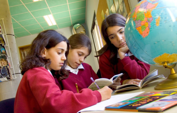 Three junior school girls studying the globe atlas for geography studies in school library_A0NP28